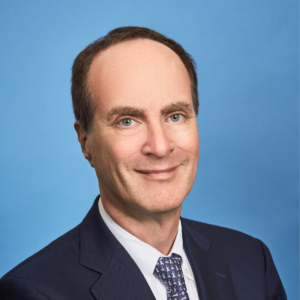 A man with short brown hair, wearing a dark suit, white shirt, and patterned tie, smiles against a plain blue background. He exudes professionalism typical of hospitality experts or those in protection services.
