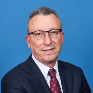 A middle-aged man with short gray hair and glasses is wearing a dark suit, white dress shirt, and a red patterned tie. He is smiling slightly and standing against a solid blue background, exuding an air of professionalism often associated with security services.