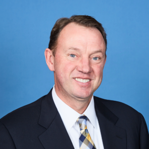 A man with short brown hair is smiling in a professional headshot. He is wearing a dark suit jacket, a white shirt, and a patterned tie. The background is solid blue, hinting at his expertise in security services.