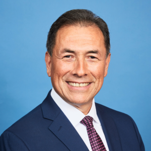 A man dressed in a navy blue suit, white shirt, and patterned tie is smiling at the camera against a plain blue background, reflecting the professionalism of top-tier Security Services.