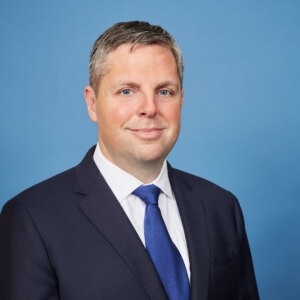 A man with short brown hair, dressed in a dark suit, white shirt, and blue tie, stands against a plain blue background. He is smiling slightly and looking directly at the camera, exuding an air of professionalism akin to that found in the hospitality industry.