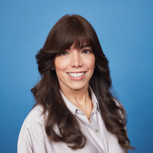 A woman with long, wavy brown hair and bangs is smiling in front of a blue background. She is wearing a light-colored button-up shirt and a gold necklace, evoking a sense of warmth and hospitality.