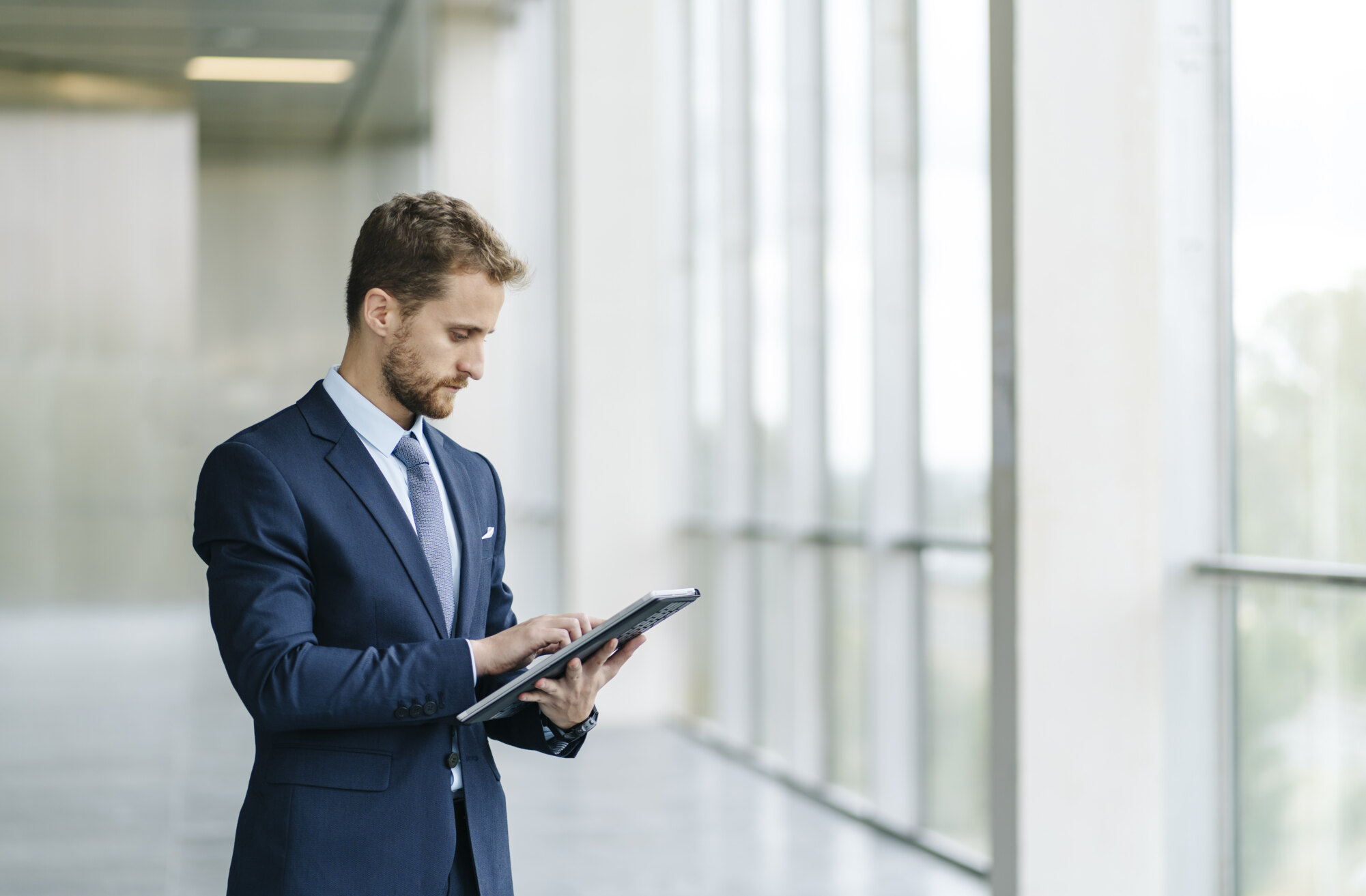 Man in a suit using a tablet near large windows in a modern office building.