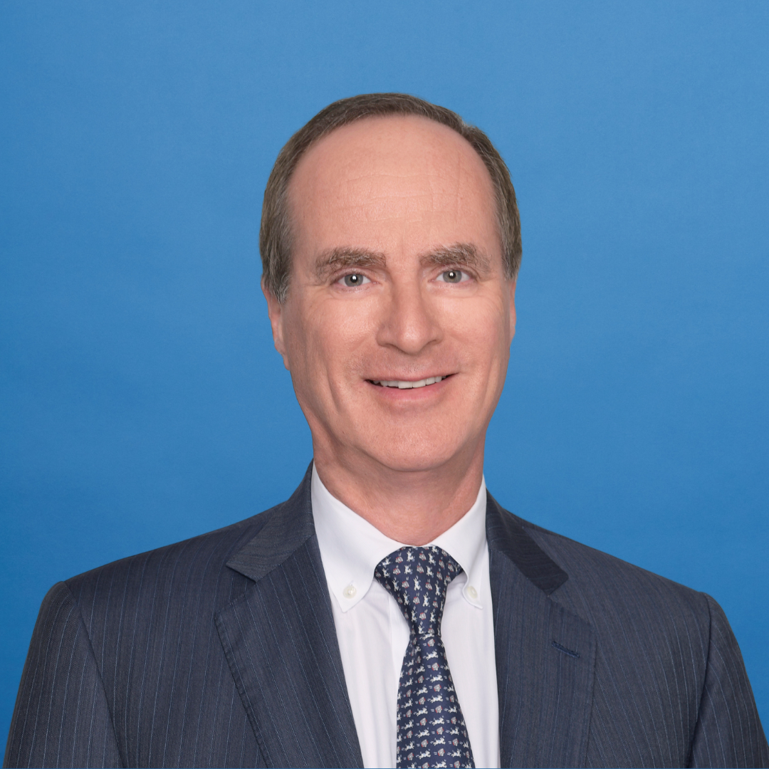 A middle-aged man in a suit and tie smiles at the camera against a plain blue background.
