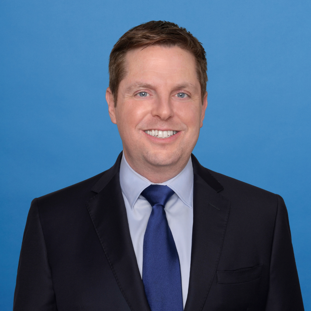 Man in dark suit and blue tie smiling in front of a plain blue background.