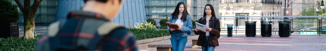 Two students walking around school campus while reading papers while another person with a backpack walks in the foreground.